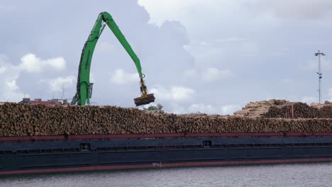 Crane-loads-timber-logs-onto-cargo-ship-full-of-timber-logs-in-sea-port,-static,-wide-shot