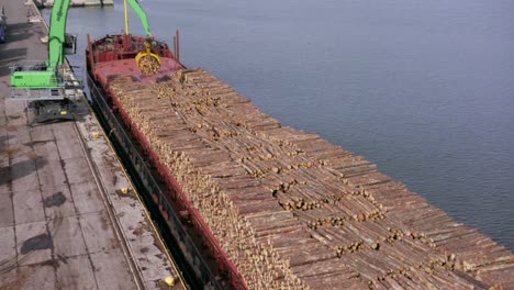 Cargo-ship-full-of-timber-logs-at-port-crane-loads-logs-from-truck-onto-ship,-slow-pan-tilt,-wide-shot
