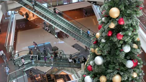 Los-Compradores-Viajan-En-Escaleras-Mecánicas-En-Un-Centro-Comercial-Mientras-La-Instalación-De-Un-árbol-De-Navidad-Se-Ve-En-Primer-Plano