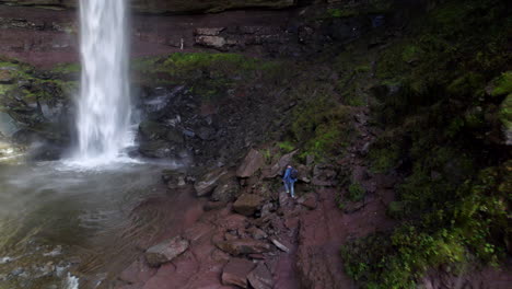 Aerial-Of-Male-Walking-Over-Rocks-Towards-Cascading-Waterfalls-In-The-Catskills,-NY