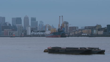 Pontoon-on-River-Thames-with-Ship,-Thames-Tidal-Barrier-and-Canary-Wharf,-Cityscape