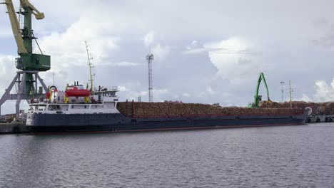 Cargo-ship-full-of-timber-logs-at-port-crane-loads-logs-onto-ship,-static,-wide-shot