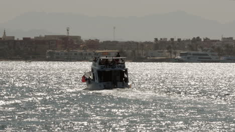 A-Yacht-Sailing-on-Sea-Towards-Coast-in-Sunny-Day-Egypt,-Sun-Glare-on-Water-Surface-and-Coastal-Cityscape-in-Horizon,-Seashore-Landscape