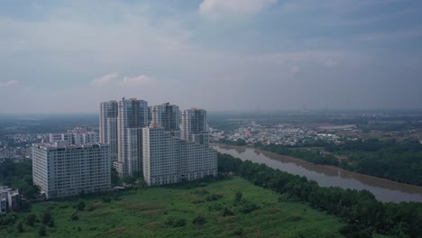 Fly-in-aerial-shot-to-large-modern-apartment-blocks-along-the-river-with-parkland-in-outer-suburbs-of-Ho-Chii-Minh-City,-Vietnam