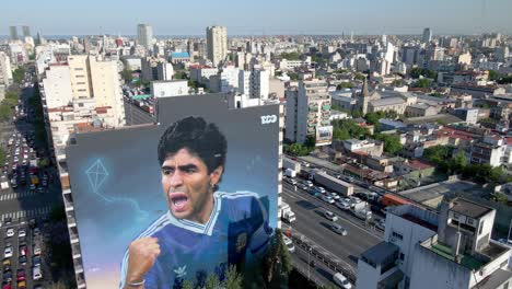 Establishing-shot-aerial-pull-out-away-from-giant-mural-of-Maradona-dedicated-to-the-soccer-legend-on-his-second-anniversary-of-death-with-downtown-cityscape-and-congested-traffics-in-the-background