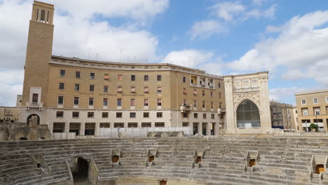 Toma-Panorámica-Del-Anfiteatro-En-Lecce,-Italia-Y-El-Instituto-Nacional-De-Seguros-En-Un-Día-Soleado