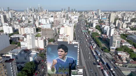 Panoramic-view,-aerial-panning-shot-of-Buenos-Aires-cityscape-with-a-giant-wall-mural-of-late-soccer-player-Diego-Maradona-on-his-anniversary-of-death,-painting-depicted-him-in-World-Cup-final-1990