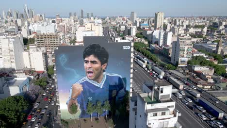 Argentines-mark-second-anniversary-of-Diego-Maradona's-death,-late-soccer-legend’s-face-brushes-the-immensity-of-the-skies-at-downtown-Buenos-Aires,-Constitución-neighborhood,-aerial-panning-shot