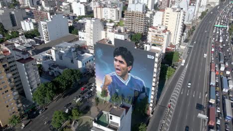 Birds-eye-view-fly-around-giant-wall-mural-of-legendary-soccer-player-Maradona-in-Constitución-neighborhood,-Buenos-Aires-at-rush-hour-with-heavy-traffics-on-Avenue-San-Juan-and-Autopista-25-de-Mayo