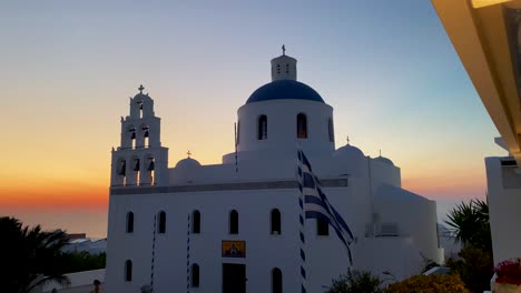 Greek-Flag-In-Front-Of-Orthodox-Church-Of-Oia-On-The-Island-Of-Santorini-In-Greece