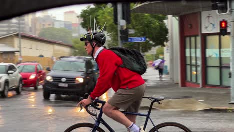 Australian-man-cycling-across-the-road-wearing-unique-design-helmet-with-cable-ties-to-deter-swooping-magpie-bird-on-a-rainy-day-at-Bowen-Hills,-Brisbane-city,-Queensland,-Australia