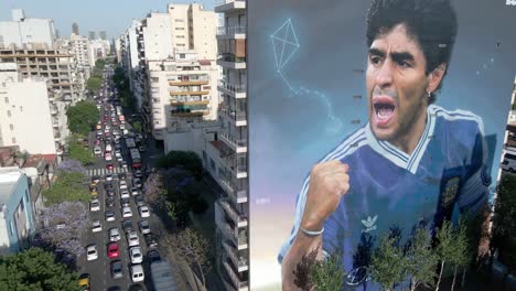Stationary-aerial-shot-capturing-a-giant-mural-of-Maradona-on-the-building-wall-with-heavy-one-way-traffics-on-San-Juan-avenue-in-the-neighborhood-of-Constitución-in-downtown-Buenos-Aires,-Argentina
