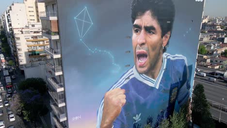 Hand-of-gold,-late-soccer-legend-Diego-Maradona-depicted-on-the-building-exterior-in-Constitucion-neighborhood,-artist-Martin-Ron-commemorate-this-mural-artwork-to-the-legendary,-aerial-panning-view