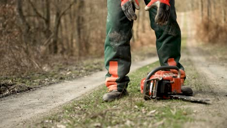 Low-Angle-Ground-Level-Shot-Of-Arborist-Picking-Up-Chainsaw-And-Walking-Away-On-Forest-Path