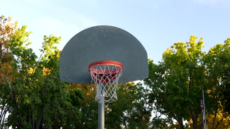 Slide-right-shot-of-a-basketball-hoop-with-metal-backboard-and-and-orange-rim-in-an-empty-park-court-at-sunrise