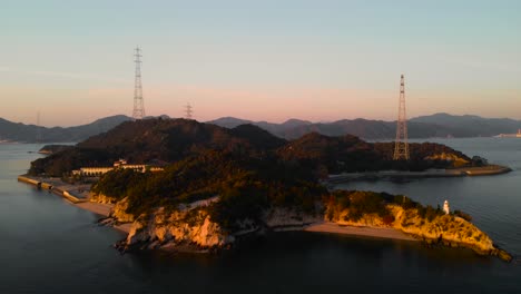 Backwards-Aerial-Drone-movement-over-island-with-lighthouse-and-powerlines-at-sunset