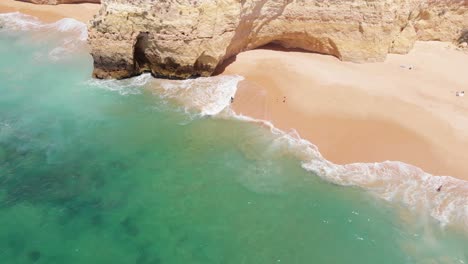 Aerial-flying-backwards-over-clear-turquoise-beach-with-rock-formation,-Algarve
