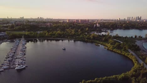Aerial-Sunset-Descending-Shot-Of-Sailboat-And-Marina-Yacht-Club-Dock-In-Lake-Bay-Surrounded-By-Green-Trees-With-City-Buildings-Skyline-In-Background-In-Toronto-Ontario-Canada