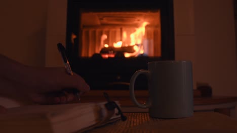 cozy-background-of-a-young-female-hand-writing-on-a-old-notebook-near-a-warm-fireplace,-with-a-cup-with-hot-steam-getting-out-of-it