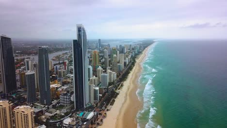 Aerial-forward-moving-view-of-skyscraper-city-by-the-beach-with-turquoise-blue-ocean-on-a-cloudy-day