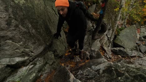 Young-Man-is-Tired-from-Climb-Up-Rocks-in-Colorful-Fall-Forest,-Handheld-pan