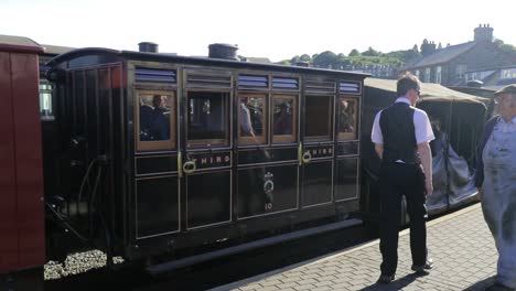 Carriages-of-tourists-and-sightseers-pull-into-Porthmadog-Harbour-Railway-Station,-Ffestiniog-Railway,-as-guard-and-engineer-cross-paths-on-platform,-engineer-pauses-to-admire,-SLOMO-60fps