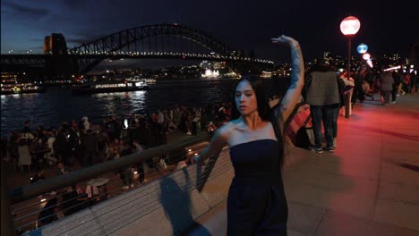 Dancing-woman-in-black-dress---dark-skinned-young-woman-with-long-flowing-hair-and-black-dress-free-dancing-in-Sydney-Opera-House-precinct-at-night,-backdrop-of-Sydney-Harbour-Bridge---4K,-59