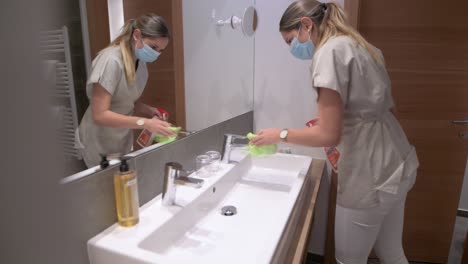 Cleaning-lady-with-face-mask-cleans-the-washbasin-in-a-hotel-room