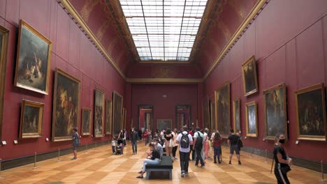 Tilting-shot-inside-a-room-with-painting-located-in-the-Louvre-Museum,-visitors-with-masks-looking-around
