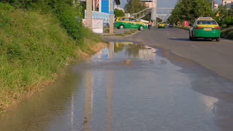 Hombre-Conduciendo-Una-Motocicleta-Conduciendo-Por-Una-Carretera-Con-Inundación-Debido-A-Una-Tubería-Rota-Subterránea-En-Tailandia