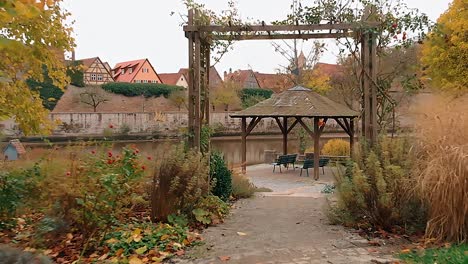 a-drone-flies-low-and-slowly-pass-through-an-wooden-arch-then-through-a-gazebo-by-the-pond-as-it-continues-to-fly-over-the-lake-and-in-front-is-a-historic-city-in-germany