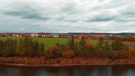 drone-flies-over-the-shore-of-a-lake-there-is-a-green-meadow-a-small-town-and-a-cloudy-blue-sky