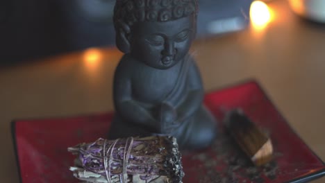Small-Buddha-statue-relaxing-with-burning-smoking-lavender-sage-sitting-on-red-rectangular-Asian-plate-on-table-shelf-in-4K-60fps