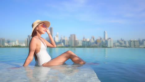 Sexy-Exotic-Woman-Sunbathing-in-Rooftop-Infinity-Pool-With-Modern-City-Skyline-in-Background,-Full-Frame