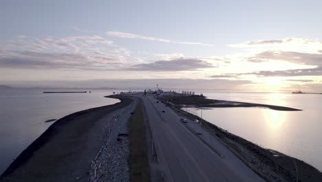Tsawwassen-ferry-port-in-British-Columbia,-drone-fly-above-highway-over-the-ocean-water-during-sunset-with-traffic-car-driving-through-the-port,-Vancouver-Canada
