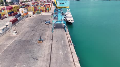 Aerial-view-of-a-woman-walking-towards-a-large-blue-crane-on-a-cargo-port-near,-pull-back-wide-shot