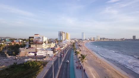 Flying-Over-Malecon-Beach-Shore-Promenade-in-Mazatlan-Mexico-Before-Sunset,-Cars-Traffic-and-People-Walking-along-Coastal-Boardwalk-Next-to-Sandy-Beach-and-Sea-Ocean,-Seaside-Cityscape