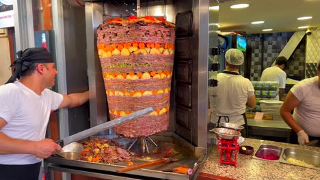 Man-cutting-meat-from-a-vertical-rotisserie-stick-at-a-kebab-restaurant-in-Bodrum-Turkey,-people-working-in-the-kitchen,-delicious-traditional-food,-seasoned-meat-with-potatoes-and-carrots,-4K-shot