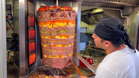 Man-cutting-meat-from-a-vertical-rotisserie-at-a-kebab-restaurant-in-Bodrum-Turkey,-people-working-in-the-kitchen,-tasty-traditional-food,-seasoned-meat-with-potatoes-and-carrots-on-a-stick,-4K-shot