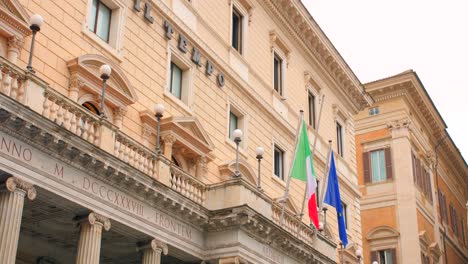 Low-angle-shot-of-Piazza-colonna-administrative-building-along-Italian-and-Euro-flag-in-Rome,-Italy-at-daytime