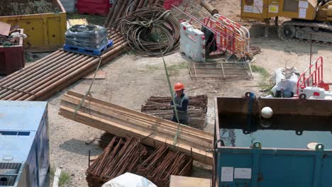 Ein-Chinesischer-Ingenieurarbeiter-Richtet-Einen-Stapel-Holzbretter-Ein,-Während-Ein-Kran-Sie-Bei-Einem-Bauentwicklungsstandortprojekt-In-Hongkong-Hochzieht