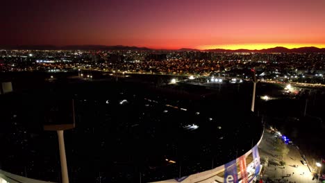 Incredible-orange-sunset-over-the-night-skyline-of-Santiago,-Chile-revealing-the-national-stadium