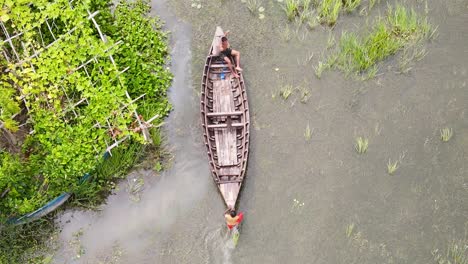 Topdown-view-follow-two-kids-on-wooden-boat-navigate-on-shallow-water,-Bangladesh