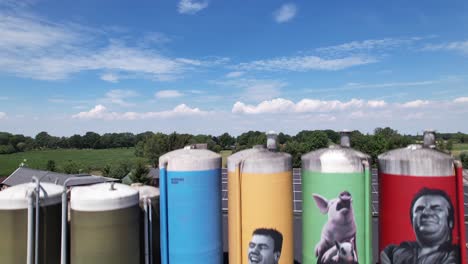 Low-aerial-approach-of-graffiti-decoration-drawing-of-farmers-family-with-animals-on-large-farm-stock-silo-containers-revealing-wider-farm-surroundings
