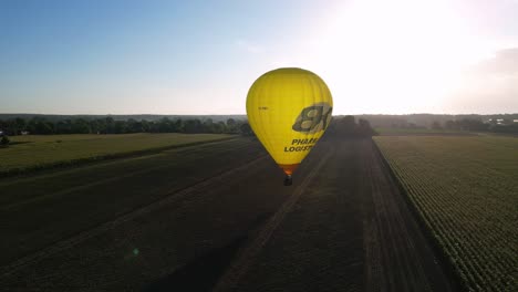 Aerial-Morning-Sunrise-View-Of-Bright-Yellow-Hot-Air-Balloon-Flying-Coming-To-Land-In-Rural-Field