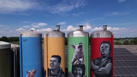Aerial-of-colorful-graffiti-decoration-drawing-of-farmers-family-with-animals-on-large-farm-stock-silo-containers-contrasted-against-a-blue-sky-with-fluffy-clouds