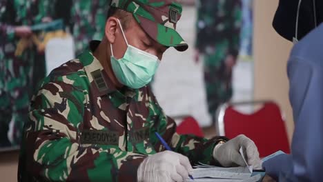 Un-Médico-Varón-De-Las-Fuerzas-Armadas-Nacionales-Indonesias-Está-Escribiendo-O-Registrando-Pacientes,-Asistido-Por-Una-Enfermera-En-El-Mostrador-De-Registro-Frente-Al-Hospital
