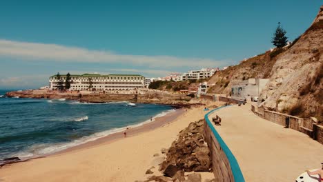 Gente-Sosteniendo-Una-Tabla-De-Surf-Caminando-En-La-Playa-De-Foz-Do-Lizandro,-Fabuloso-Resort-En-El-Fondo,-Portugal