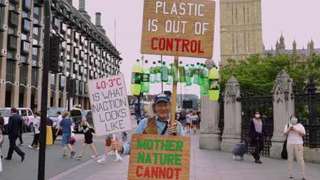 Old-male-climate-activist-in-Westminster,-London,-holding-up-climate-change-and-anti-plastic-signs-to-protest-against-CO2-pollution-and-the-climate-crisis-and-for-nature-protection-and-preservation