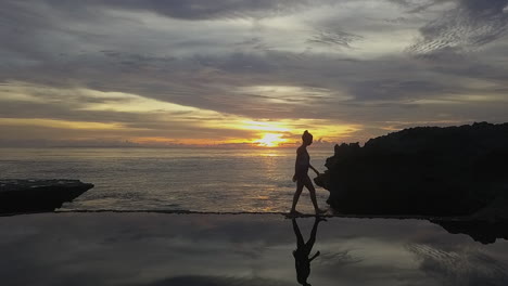 Slo-mo-woman-reflected-in-natural-pool,-walks-across-ocean-sunset